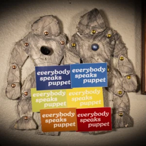 Two grey-colored rock monster puppets made of foam board with shiny metal joints hold 6 rectangular stickers in blue, green, yellow, orange, and red that read 'everybody speaks puppet'
