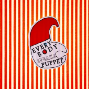A white sticker shaped like Mr. Punch with a red hat. Written on his face is 'Everybody Speaks Puppet'. The background is a red and white striped cloth.